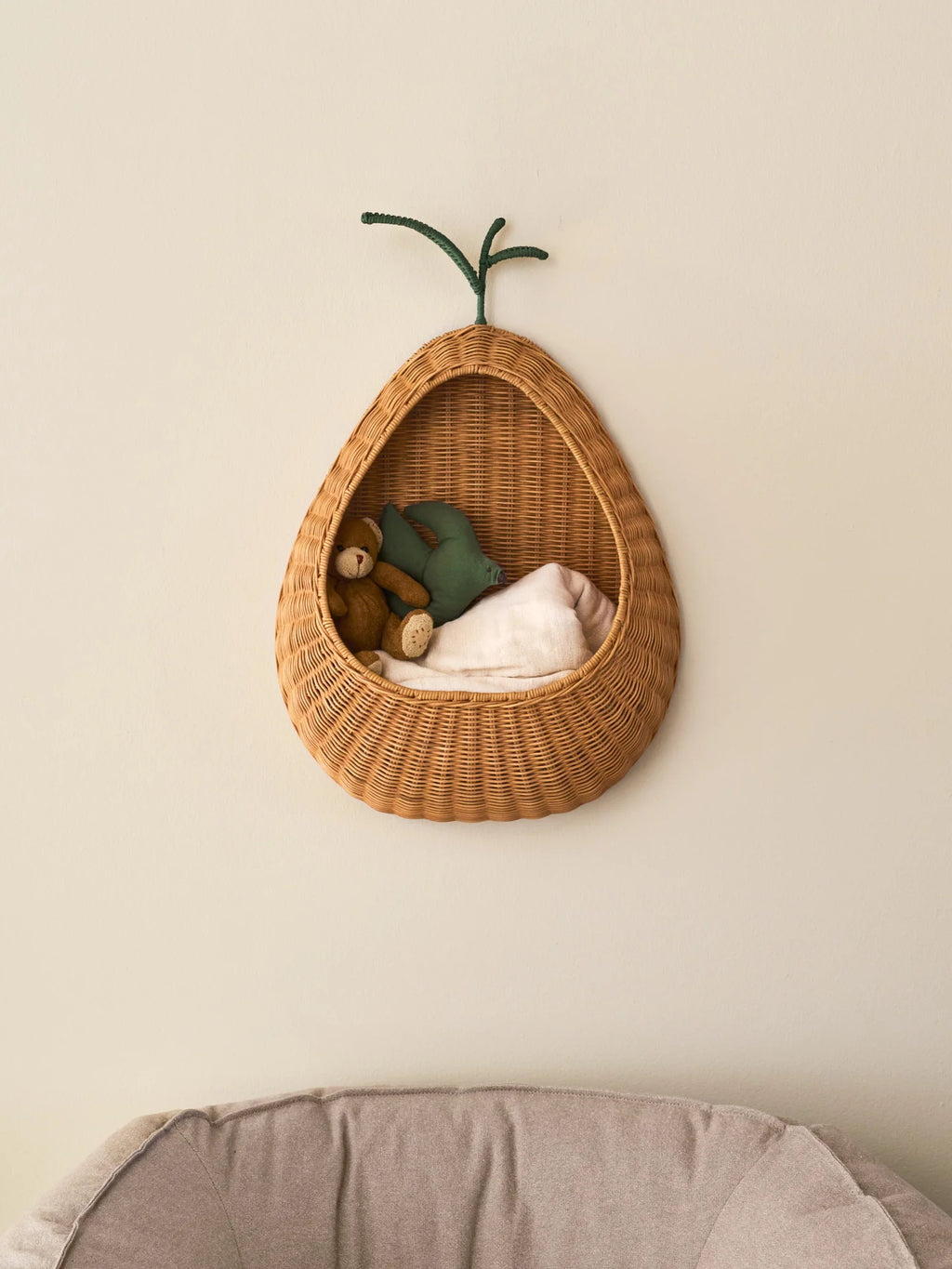 A wall-mounted pear-shaped braided rattan basket with a green leaf detail, containing various items like books and toys, set against a neutral background.