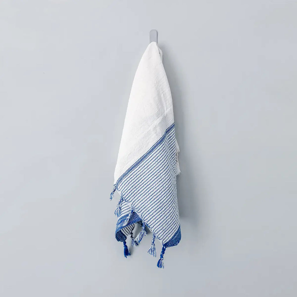 White towel with blue stripes and tassels hanging on a gray wall.
