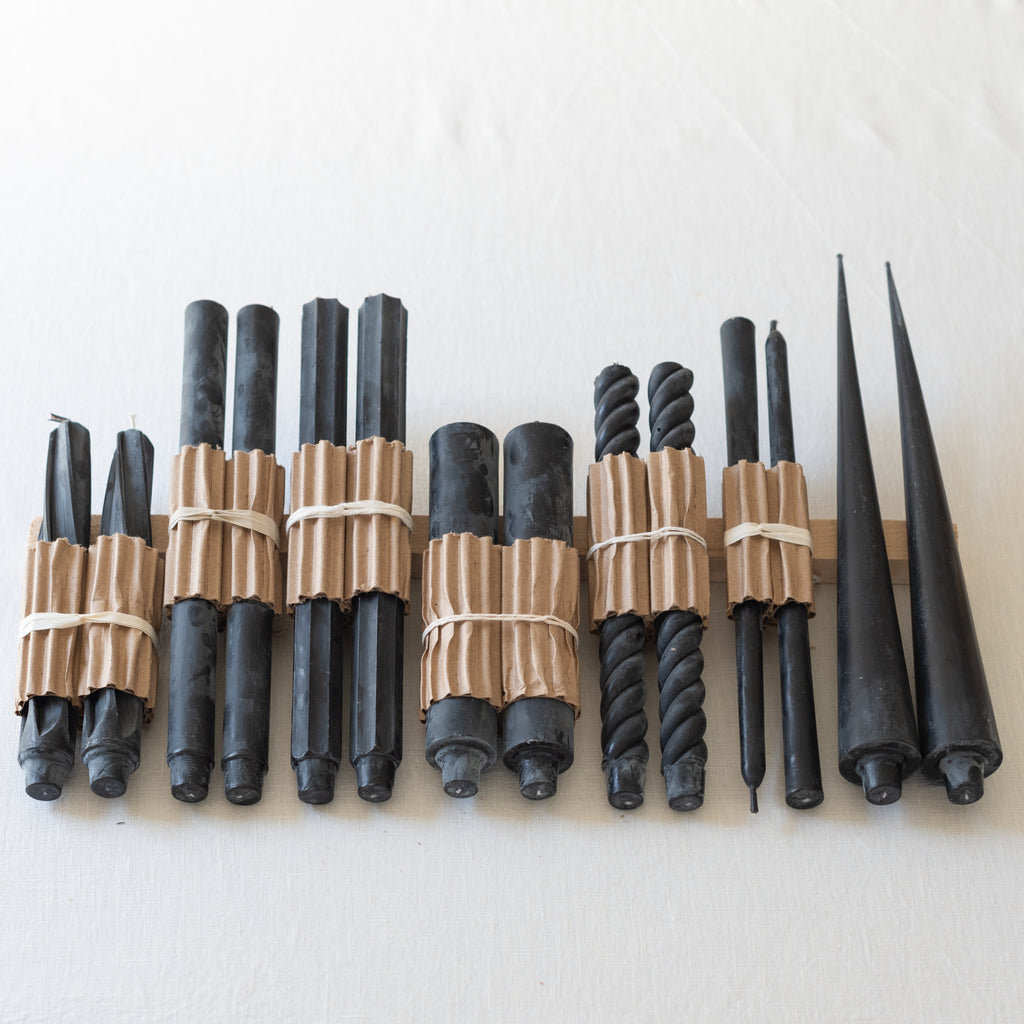 A selection of black taper candles arranged by design, with natural and twisted patterns, made from beeswax, displayed against a wooden backdrop.