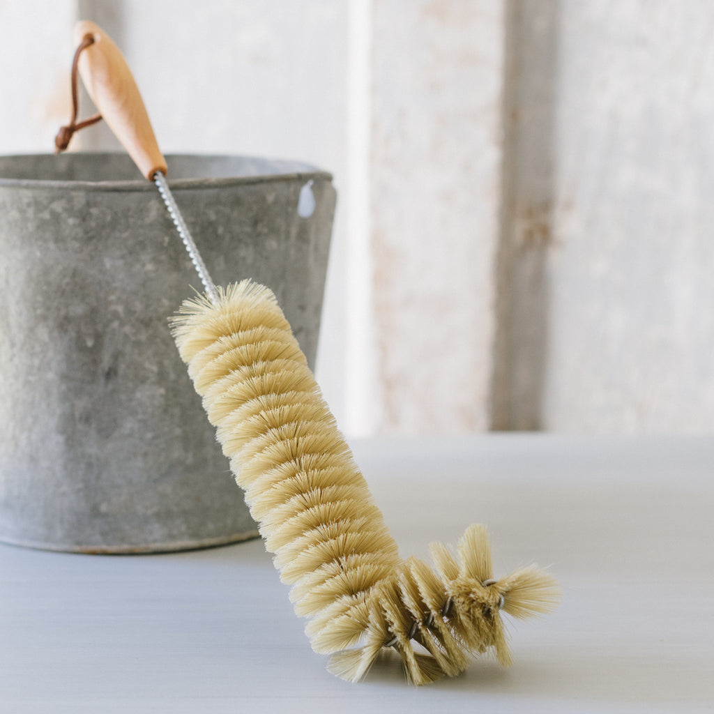 A beige radiator brush with a wooden handle and bristles, resting against a gray metal bucket on a white surface.