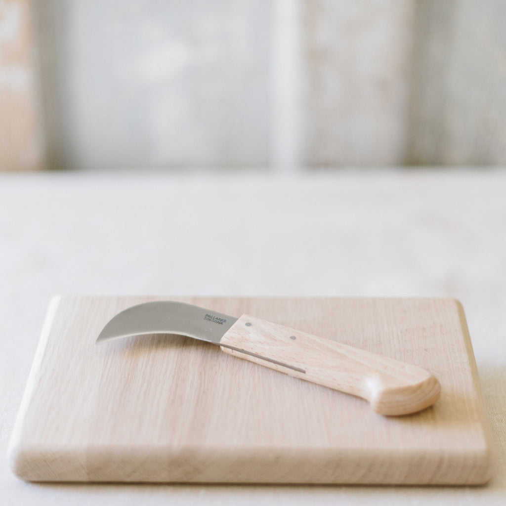 A curved carbon steel harvest knife with a beechwood handle resting on a wooden cutting board.