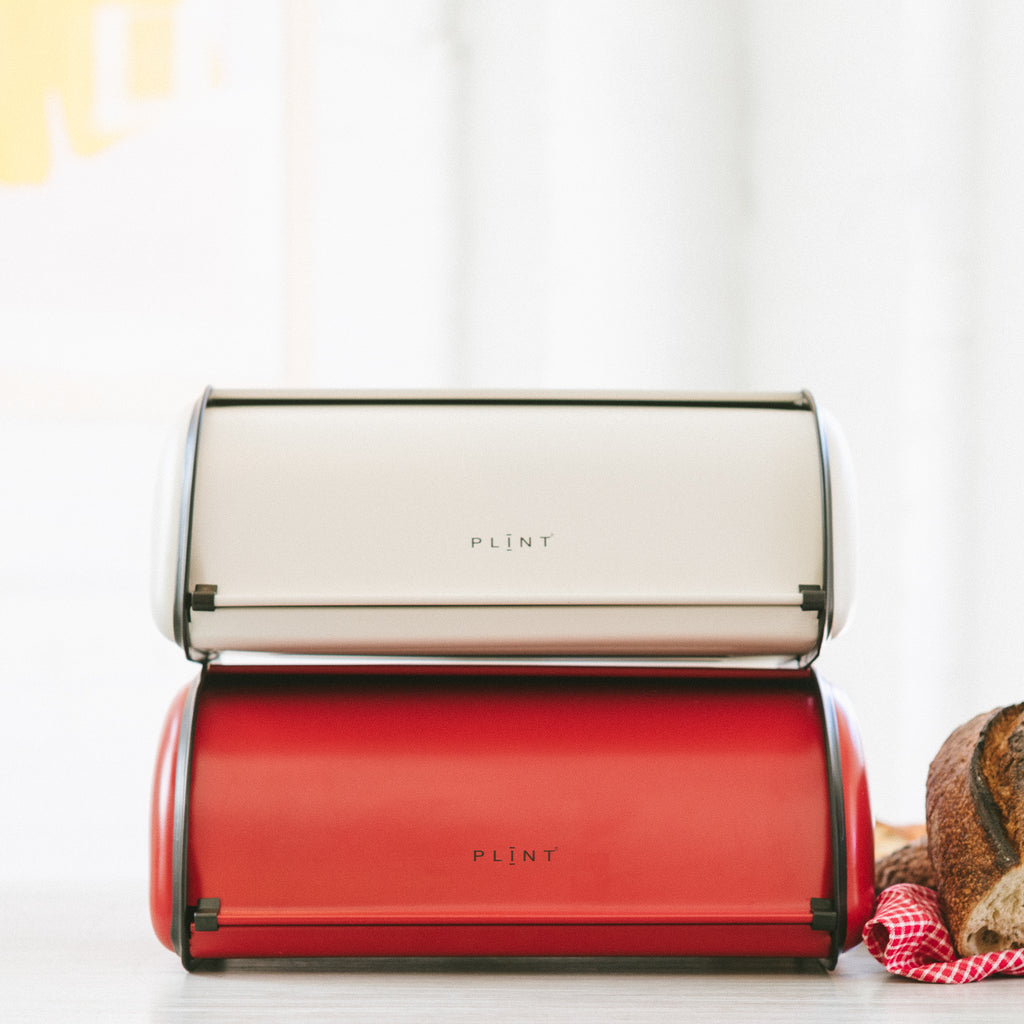 Red and cream Scandinavian-style metal bread boxes stacked on top of each other, with the brand name 'PLINT' on them, closed and placed on a kitchen counter next to some bread.
