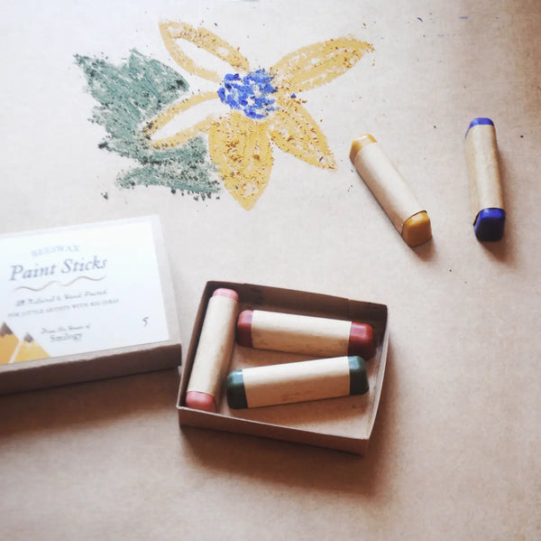A set of pastel paint sticks in a cardboard box with one open box and two loose sticks beside it, arranged on a beige surface with a flower drawing made from the sticks above.