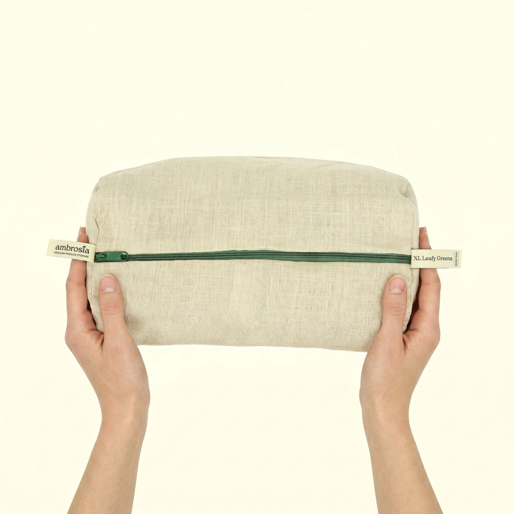 A person's hands holding a rectangular beige linen pouch with a green zipper and cream-colored pull tabs that read 'ambrosia