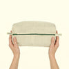 A person's hands holding a rectangular beige linen pouch with a green zipper and cream-colored pull tabs that read 'ambrosia