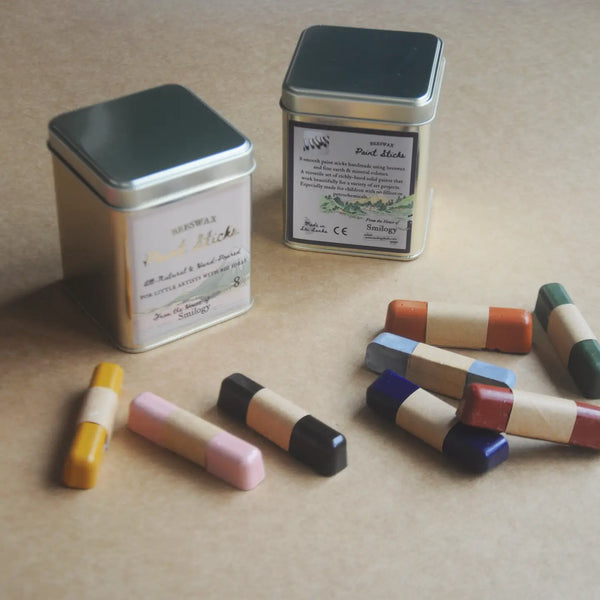 Two metal tin candles with silver lids and cream labels are displayed on a beige surface, alongside several small wooden stick candles in assorted colors including yellow, pink, black, blue, red, and green.