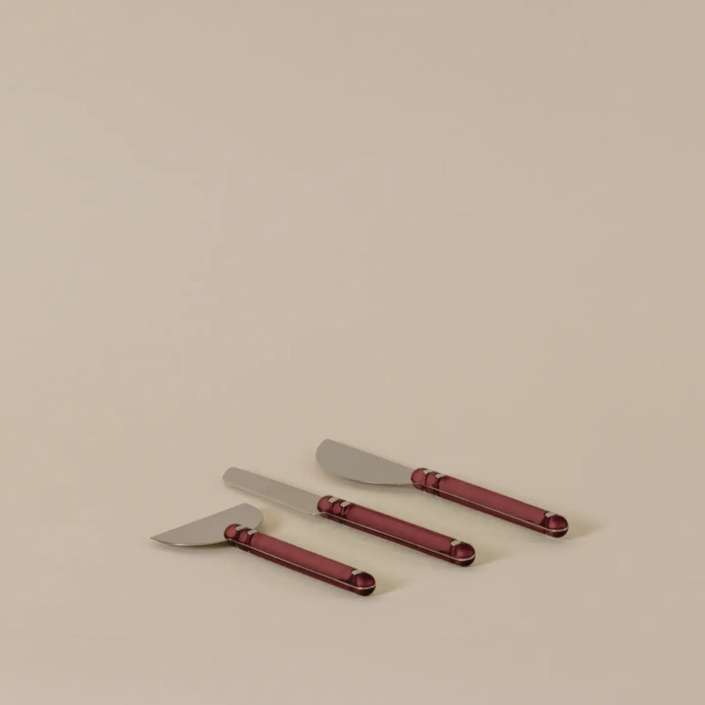 Three cheese knives with metallic silver blades and dark red handles, arranged diagonally on a plain beige background.