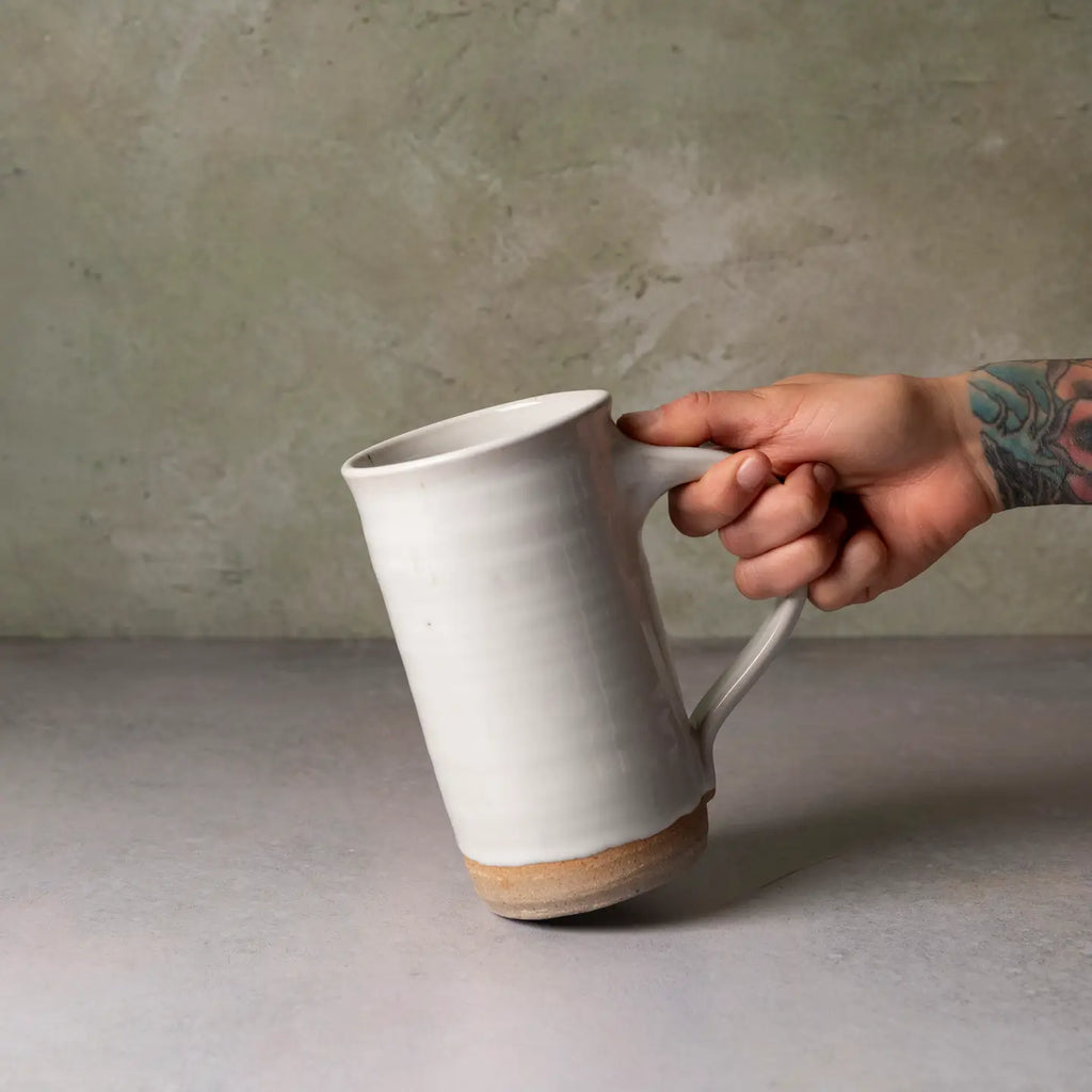 A hand with a floral tattoo holds a tall, rustic ceramic mug with a white glazed finish and a natural clay base, set against a textured gray concrete background.