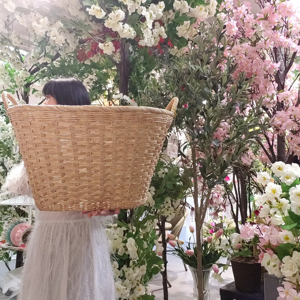 A person in a white dress carries a large natural woven wicker basket in a floral shop filled with pink and white artificial flowers.