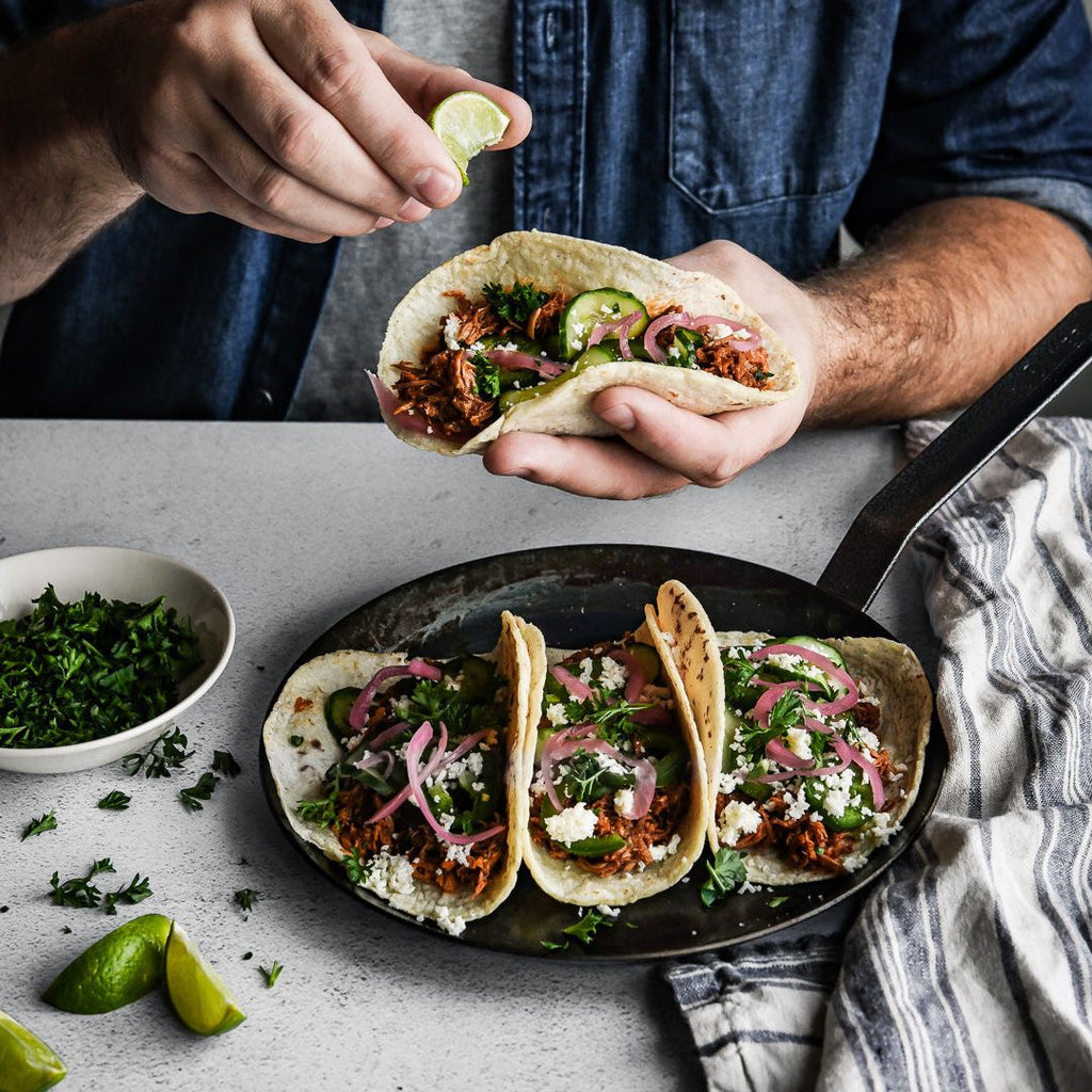 Person holding a taco with a pan of more tacos on a table.
