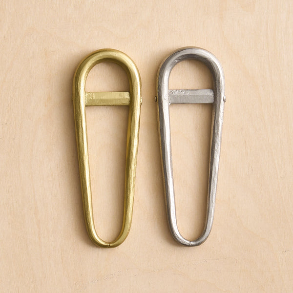 Two bottle openers, one in polished brass and one in polished silver, displayed side-by-side on a light beige wood background.