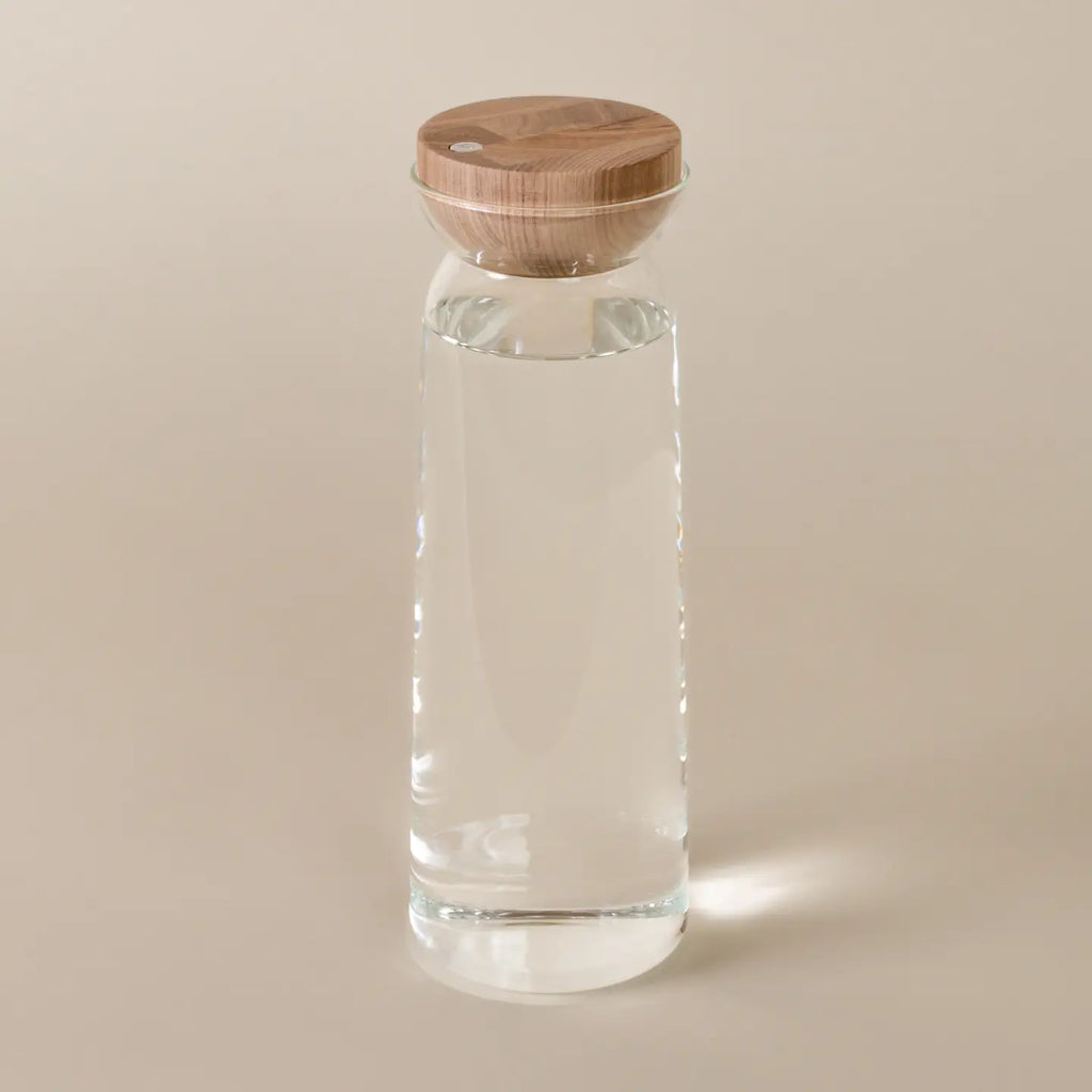 A clear glass water bottle with a round light brown wooden lid, shown on a solid beige background.