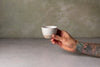 A hand with a colorful tattoo holds a small handmade ceramic cup against a textured gray concrete background.