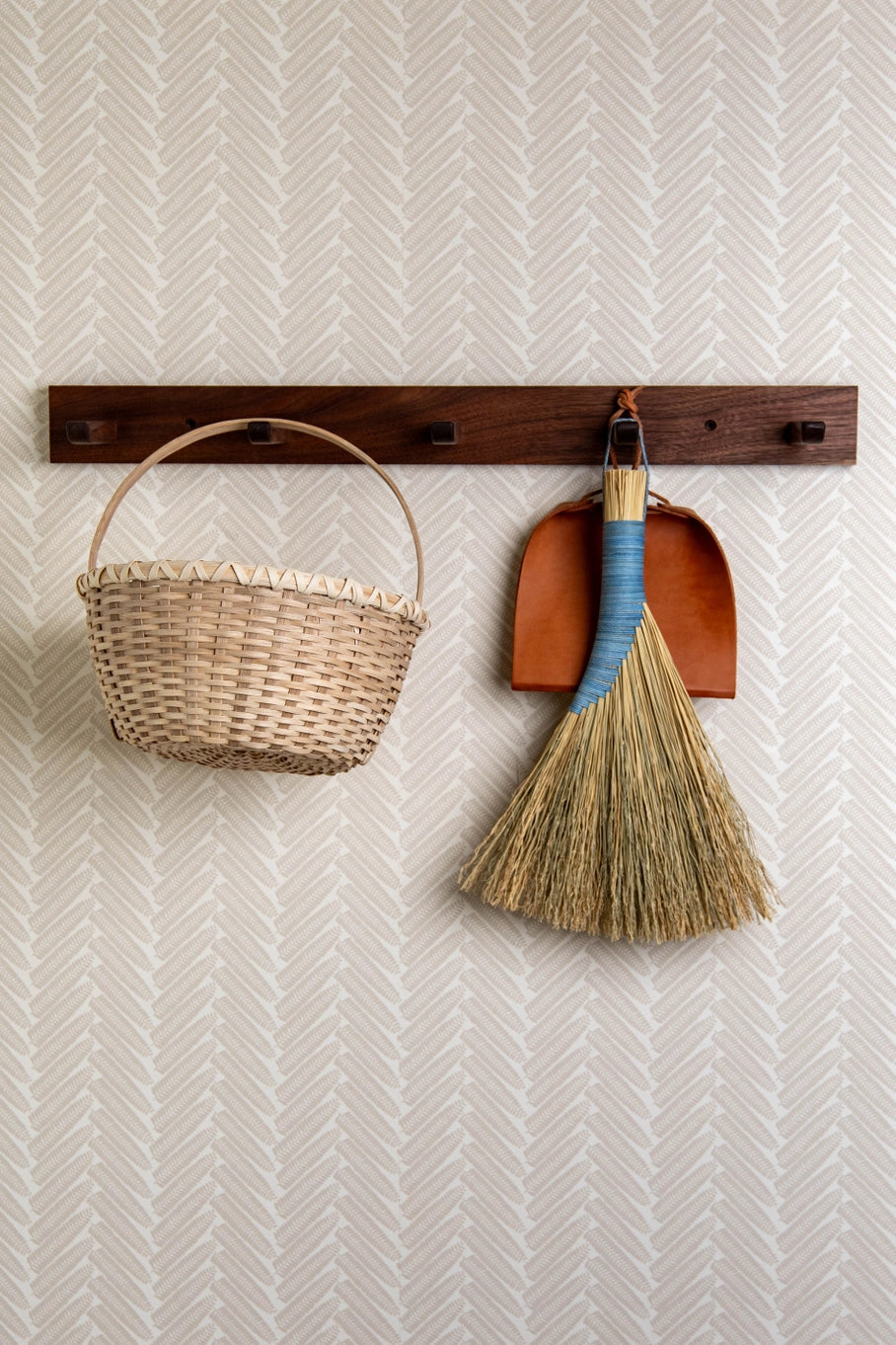 Wicker basket, broom, and dustpan hanging on a wooden rack against a white wall with a subtle pattern.