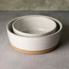 A set of two nested ceramic bowls with a speckled off-white glaze and a natural tan clay base, photographed on a textured gray surface.