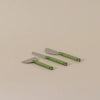 Three cheese knives with silver metal heads and translucent green handles, arranged diagonally on a plain beige background.