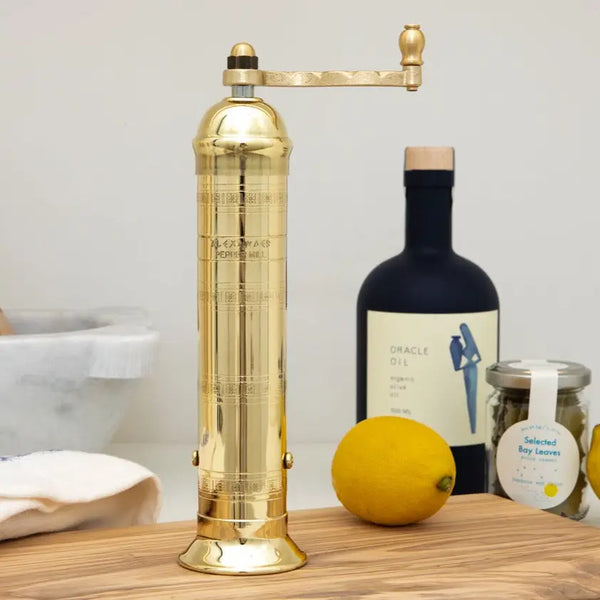 A brass manual pepper grinder with engraved markings stands on a wooden table next to a bottle of Oracle Oil, a jar of lemon thyme, and a lemon.