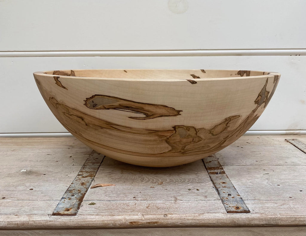 A large handcrafted wooden bowl with natural light tan wood and dark brown grain patterns, displayed on a rustic weathered wooden surface against a white paneled wall.