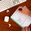 Person writing in a notebook with a keyboard, earbuds on a desk.