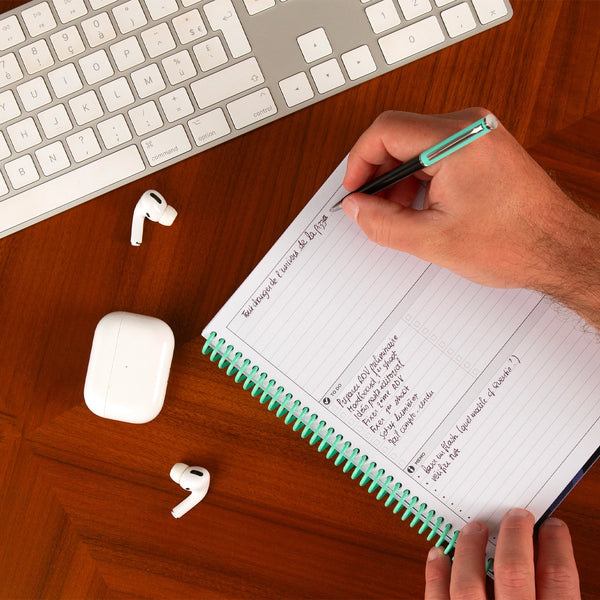 Person writing in a notebook with a keyboard, earbuds on a desk.