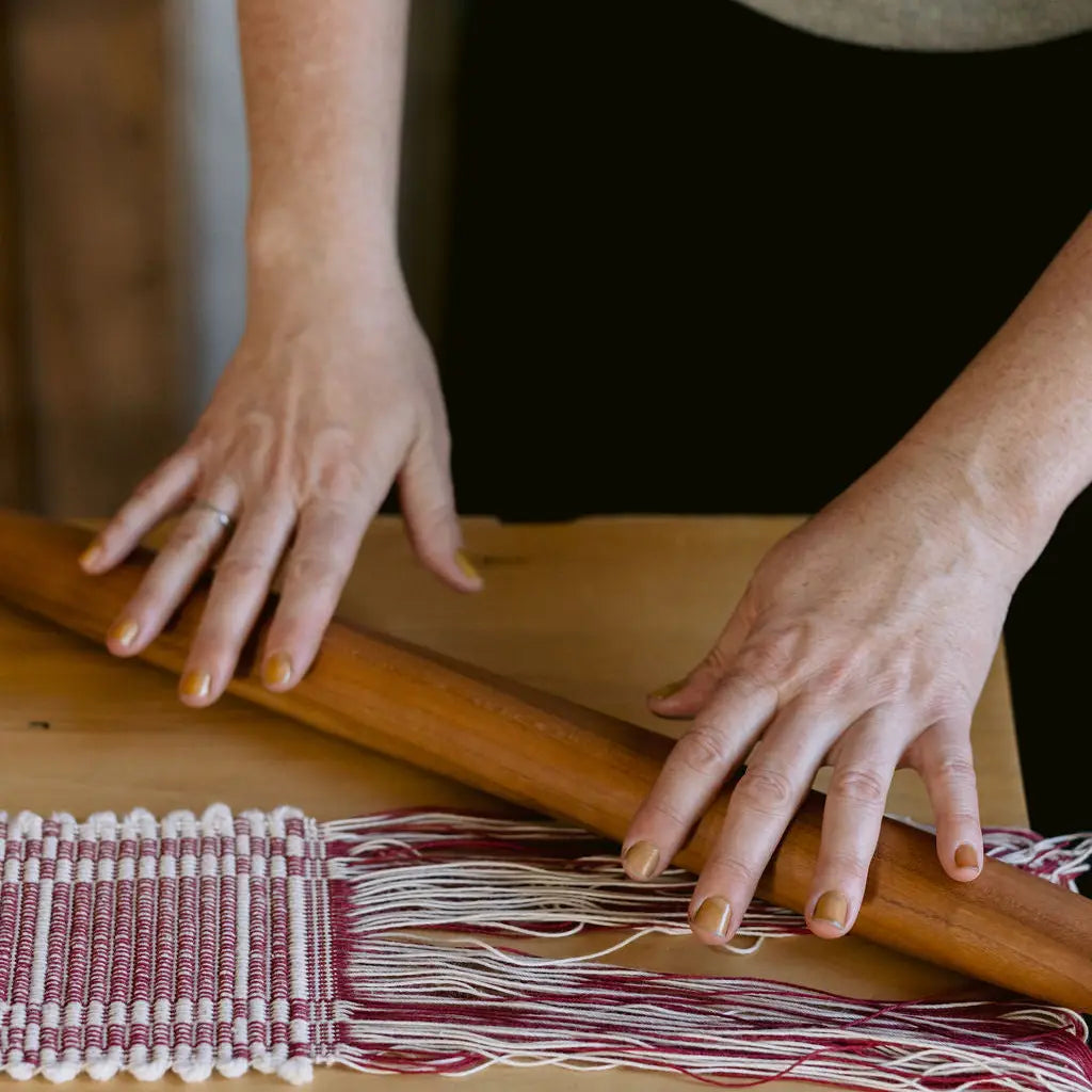 Close-up of a person's hands weaving a textile with burgundy and white threads over a wooden loom, with a wooden rolling pin resting across the woven area.