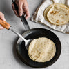 Person cooking a tortilla in a carbon steel skillet on a light gray surface with stacked tortillas nearby.