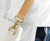 A close-up of a light tan leather shoulder strap with gold-tone hardware attached to a white canvas bag, styled over a white button-down shirt with a portion of blue jeans visible at the corner.