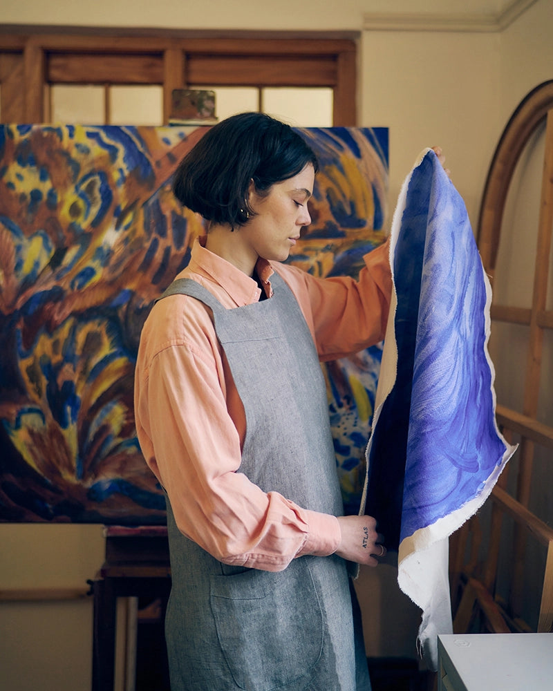 A woman in an artist's studio wearing a gray apron over a peach shirt, holding up a large blue and white abstract painting to show its full size and design. A larger, finished abstract painting in blue, yellow, and brown hangs on the easel behind her.