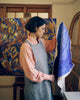 A woman in an artist's studio wearing a gray apron over a peach shirt, holding up a large blue and white abstract painting to show its full size and design. A larger, finished abstract painting in blue, yellow, and brown hangs on the easel behind her.