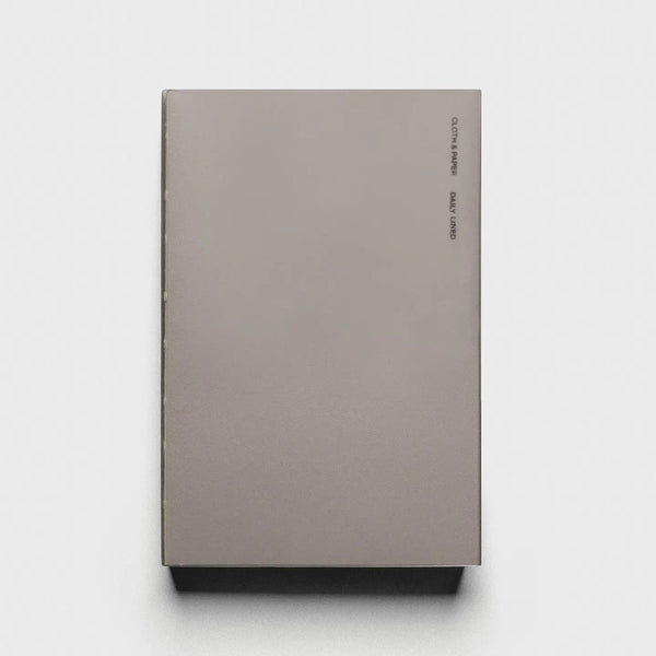 A minimalist hardcover notebook or journal in a muted taupe-gray color, featuring small black text printed vertically on the right side of the cover, set against a clean white background.