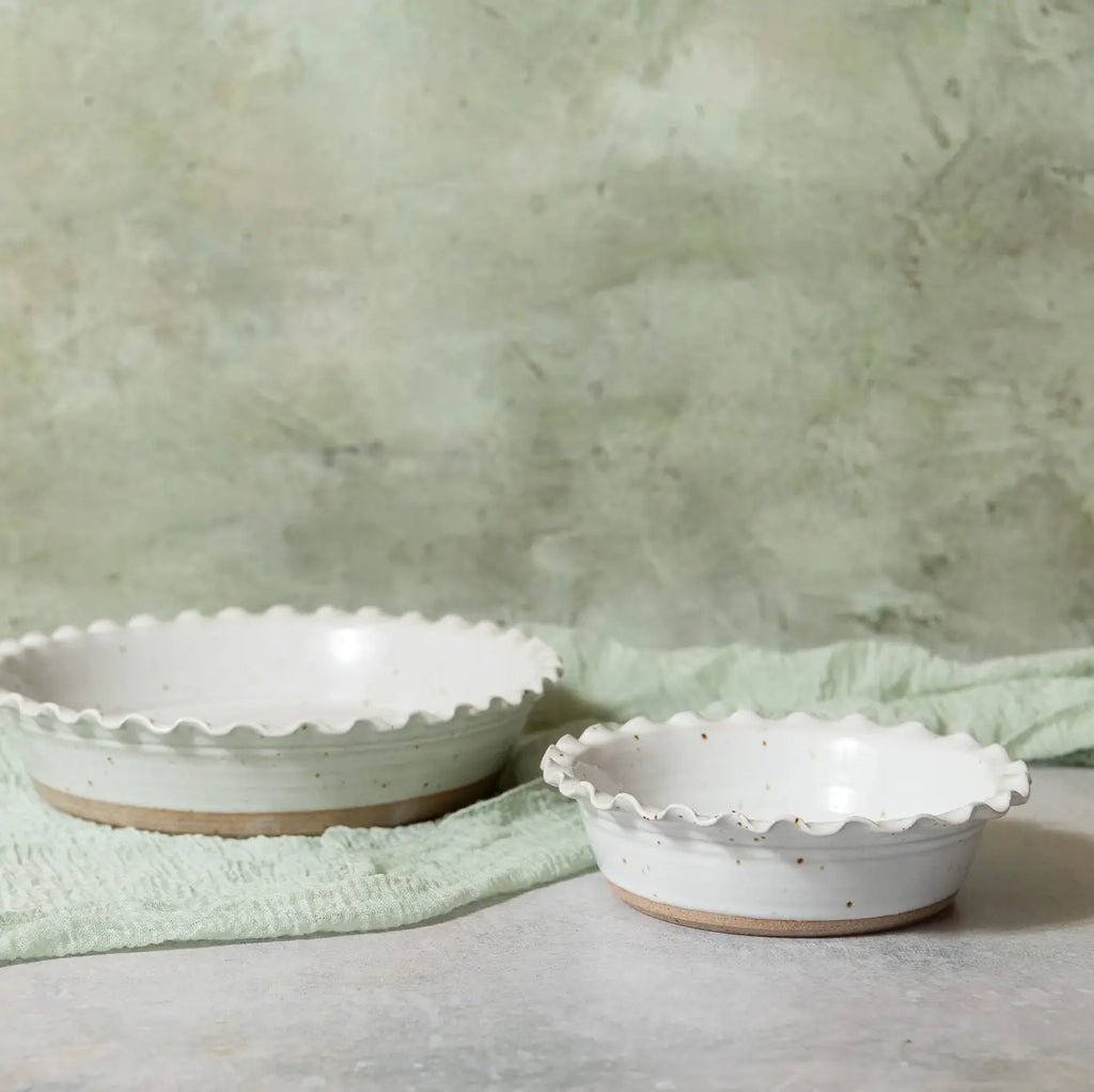Two round white ceramic pie bowls with scalloped edges and a speckled finish, displayed on a light gray surface with a pale green cloth and textured wall background.
