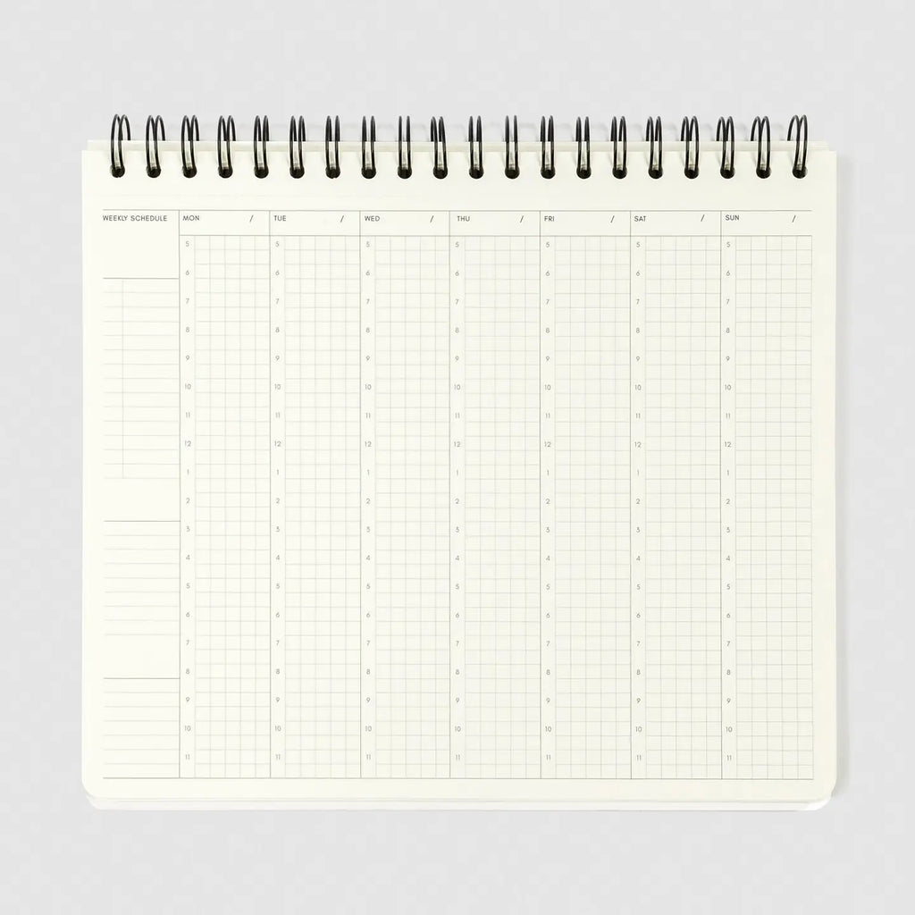 A weekly schedule planner page from an open spiral-bound notebook, showing a grid layout for Monday through Sunday with columns for days and blank spaces on the left, photographed on a light gray background.