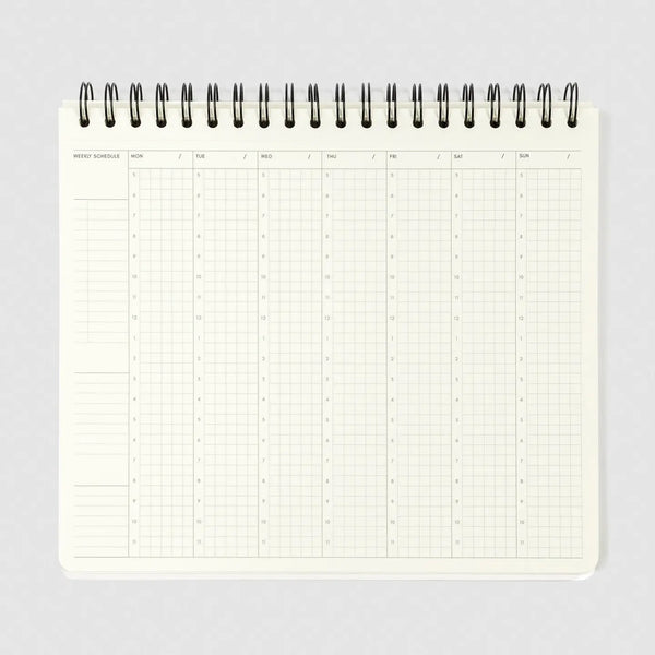 A weekly schedule planner page from an open spiral-bound notebook, showing a grid layout for Monday through Sunday with columns for days and blank spaces on the left, photographed on a light gray background.
