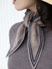 A close-up of a woman wearing a knitted scarf tied around her neck. The scarf is a warm taupe-beige color with dark navy blue stripes, paired with a gray knit top and a straw hat visible at the top edge.