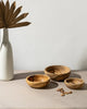 Three wooden bowls on a neutral surface with a white vase and dried leaves.