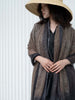 A woman wearing a wide-brimmed straw hat and a textured brown and gray shawl over a black top, standing against a white slatted wall.