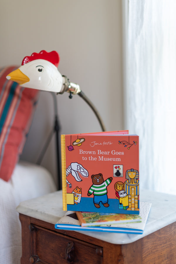 A children's picture book titled 'The Brown Bear Goes to the Museum' stands open on a small wooden table next to a decorative lamp shaped like a white rooster with a red comb and yellow beak.