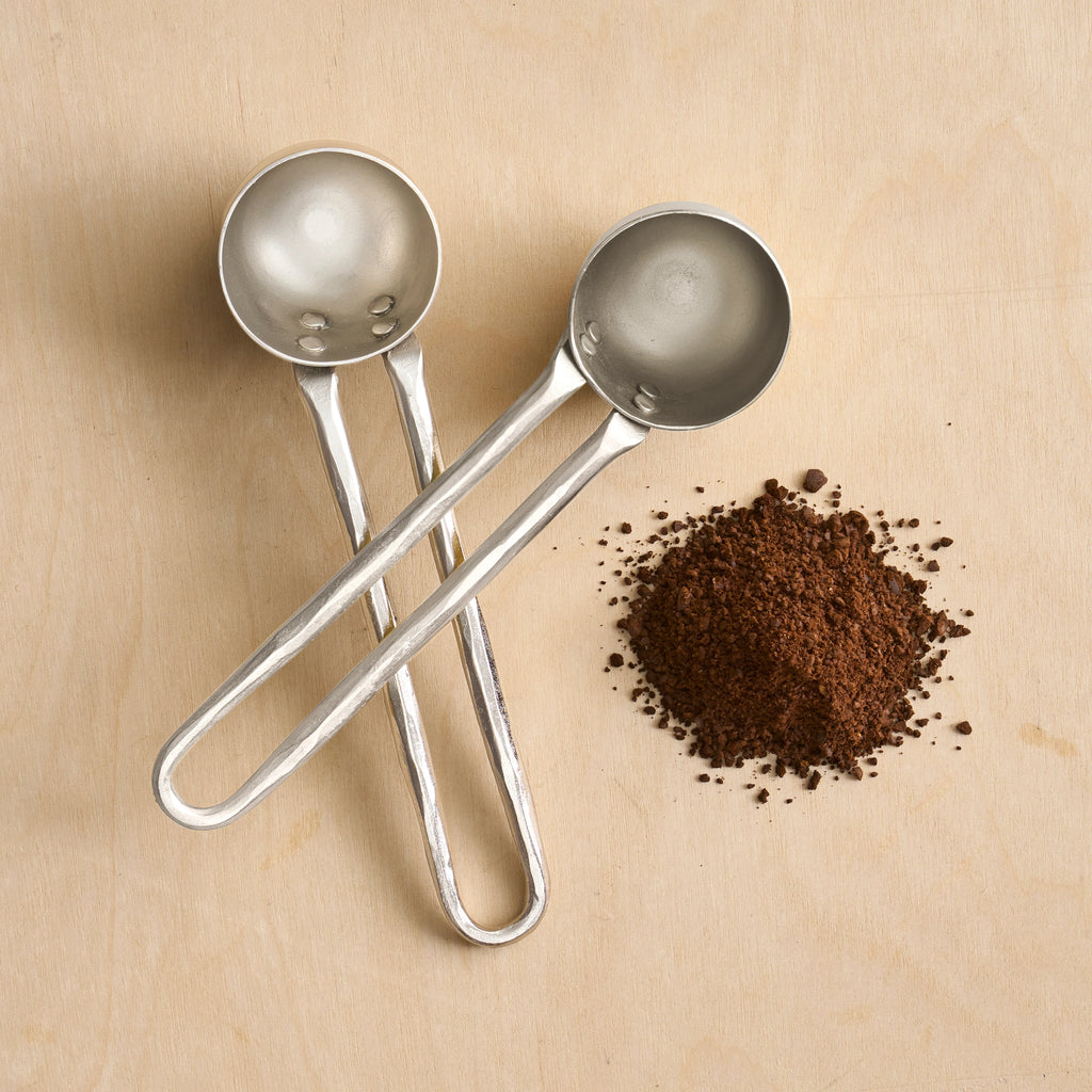 Two metal coffee scoops on a beige surface with ground coffee.