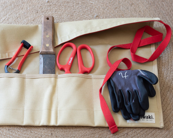 Beige tool roll with red straps, knife, scissors, and black gloves on a beige surface