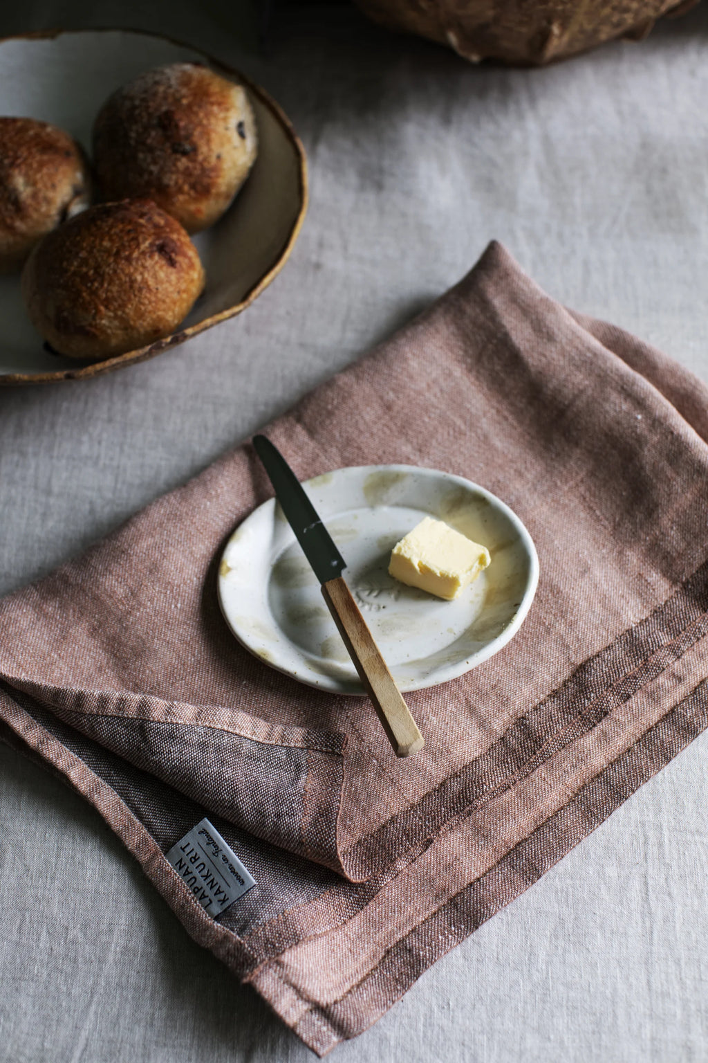 Folded kitchen towel with a small butter plate and knife on top, in the background plate with buns 