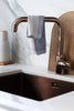 Dishtowel hanging from a kitchen faucet over a kitchen sink. A wooden cutting board behind the faucet.