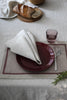 A table setting with linen placemat, tableware and linen napkin. in the background a piece of bread on a wooden cutting board. 