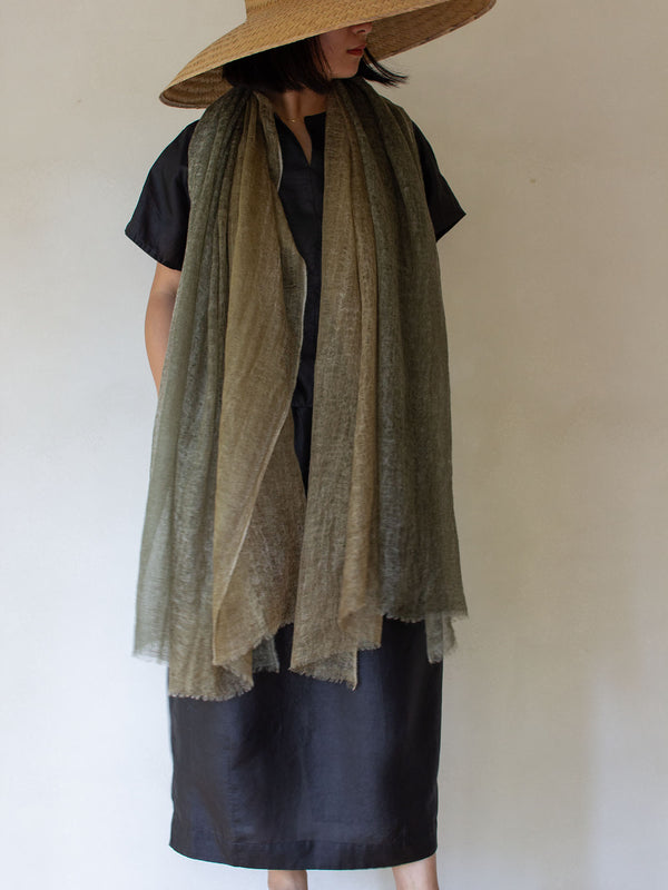 A person wearing a wide-brim straw hat, a black short-sleeve top, and a black skirt, with a long sheer olive-brown scarf draped over the shoulders against a plain light wall.