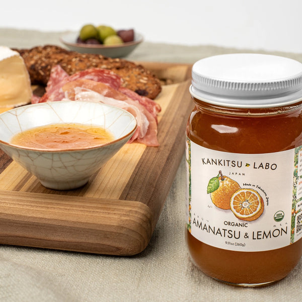 Jar of Kankitsu Labo Amanatsu & Lemon spread on a table with a bowl of soup and a plate of food in the background.