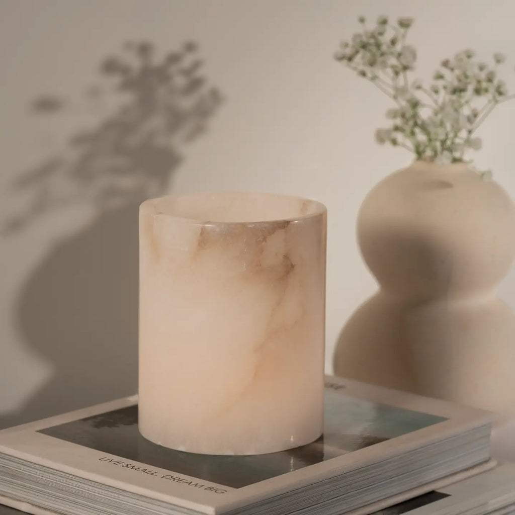A pale blush pink marble candle holder or candle vessel with soft beige veining, styled on top of books with a blurred cream vase of small flowers in the background.