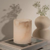 A pale blush pink marble candle holder or candle vessel with soft beige veining, styled on top of books with a blurred cream vase of small flowers in the background.