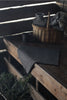 A dark charcoal gray wool seat pad is draped over a rustic wooden bench, with a matching gray throw and a wicker basket holding a dark vase in the background.