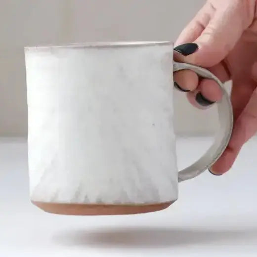 A hand with black nail polish holds a handmade ceramic mug with a textured off-white glaze and a terracotta-colored base against a neutral light background.