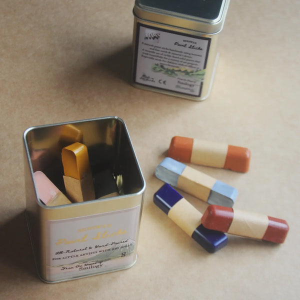 A tin of pastel sticks on a beige background, with one open tin showing the chalk inside and several loose chalk sticks arranged beside it.