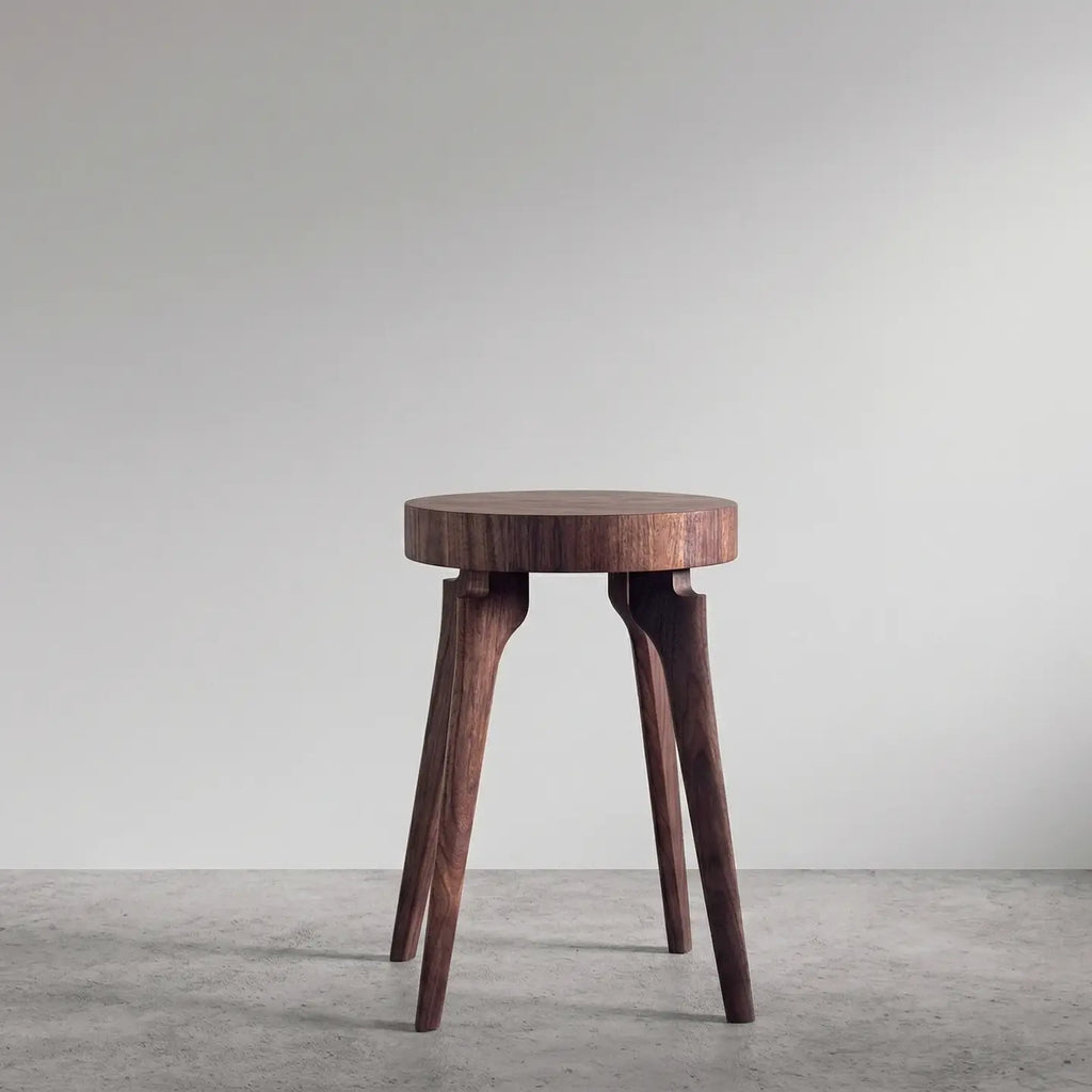 A dark brown wooden side table with four tapered legs, photographed in a minimalist studio setting with a light gray wall and concrete floor.